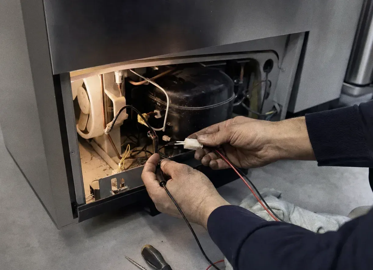 Lower rear panel removed from a refrigeration cabinet, exposing the compressor and wiring with meter probes on the terminals